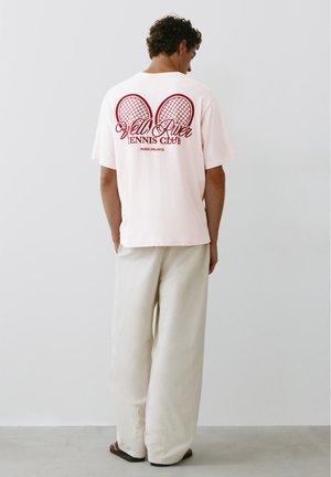 Uomo che indossa una maglietta rosa pallido con racchette da tennis rosse e testo "Vell River Tennis Club Paris France" sulla schiena, pantaloni crema e sandali scuri.