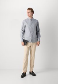 Ανοιχτό μπλε πουκάμισο με κουμπιά σε ελαφρύ ύφασμα, συνδυασμένο με μπεζ παντελόνι. Το άτομο κρατάει μια μαύρη ταμπλέτα και φοράει μαύρα παπούτσια.