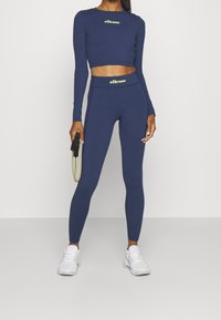 Top corto de manga larga en color navy y leggings de tiro alto a juego con acentos de logo amarillo, combinados con zapatillas deportivas blancas y una cuerda para saltar.