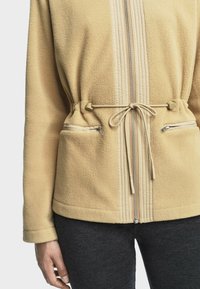 Veste en polaire de couleur beige clair, avec une fermeture éclair à l'avant, une taille à cordon de serrage et deux poches zippées. Tissu texturé avec un accent lisse.
