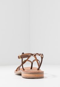 Sandale en cuir marron avec des sangles tressées et une boucle dorée. Semelle en bois plate avec un design superposé. Style simple et ouvert au niveau des orteils.
