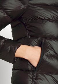 Main d'une personne insérée dans la poche zippée d'une doudoune matelassée noire avec des coutures visibles.