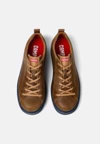 Sneaker in pelle marrone con lacci beige, soletta rossa e suola in gomma blu navy. Presentano dettagli forati sulla punta e un'asola sul tallone.