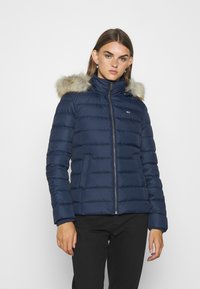 Chaqueta puffer azul marino con cierre de cremallera, que presenta una capucha con borde de piel sintética, puños acanalados y costuras horizontales en toda su superficie.