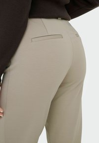 Gros plan sur un pantalon beige sur mesure avec des poches à passepoil à l'arrière, porté avec un haut marron foncé.