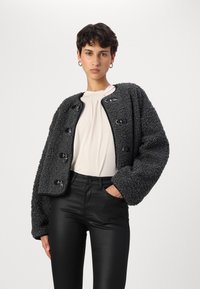 Giacca nera strutturata con un design ricamato e soffice, dotata di bottoni lucidi a contrasto e maniche larghe, abbinata a pantaloni neri.