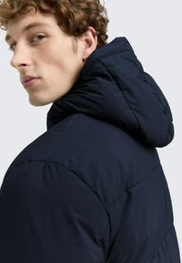 Giacca puffer blu navy con design imbottito e cappuccio. Presenta una texture liscia e dettagli cuciti lungo le spalle e le maniche.