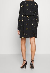 Robe noire à manches longues avec une taille cintrée, ornée d'un motif de petites fleurs jaunes. Tissu lisse ; associée à des talons noirs.