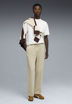 Pantalones de chándal beige y una camiseta blanca con bolsillo, combinados con un suéter a rayas beige claro y marrón. Gafas de sol amarillas completan el look.