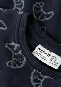 Ciemnoniebieska bluza z nadrukiem szarych łapek kota, z ryflem przy kołnierzu oraz metką z rozmiarem i instrukcjami pielęgnacji. Wykonana z bawełny.