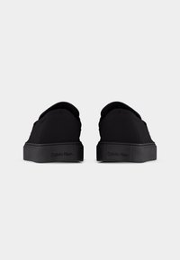 Mustade madala säärega tossude paar, millel on paksud tallad ja Calvin Kleini logo tagumistel kandadel, vaadates tagant valgel taustal.