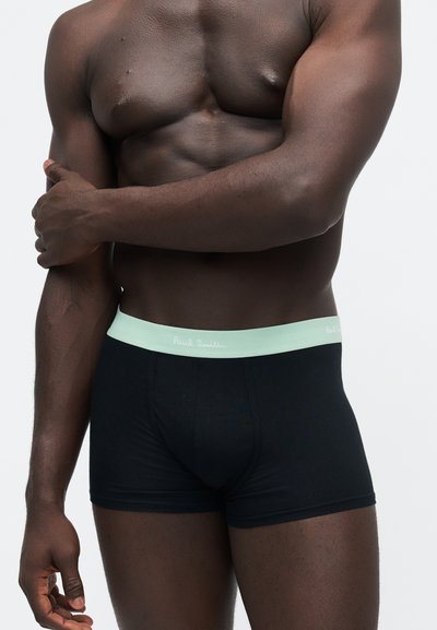 Homme musclé à la peau foncée portant un boxer noir avec une ceinture vert pâle, debout un bras croisé sur la poitrine.