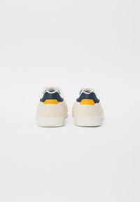 Zapatillas blancas con un talón de ante crema texturizado, acento navy en la parte trasera y un detalle en naranja brillante. Parte delantera lisa con una suela de goma.