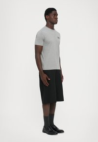 Mann som står sidelengs iført en grå EA7 t-skjorte, svarte lange shorts, mørke sokker og svarte skinnsko mot en enkel hvit bakgrunn.