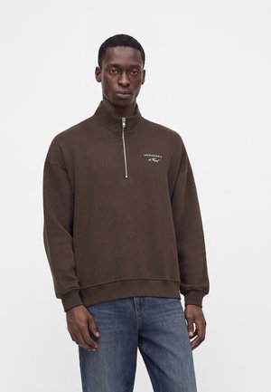 Mężczyzna w ciemnobrązowej bluzie z krótkim zamkiem i logo Abercrombie oraz niebieskich dżinsach stoi na tle gładkiego białego tła.