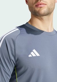 T-shirt de sport gris à col rond, avec un design à trois bandes blanches sur les épaules et un logo. Tissu lisse avec un léger éclat et des surpiqûres accentuées.