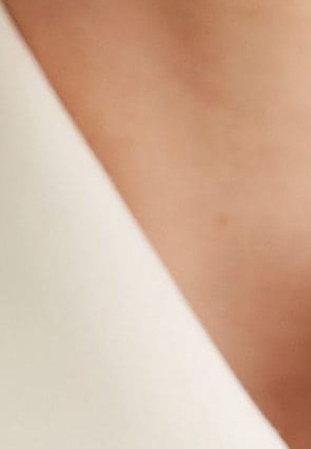 Close-up van soepel, lichtbeige stof met een zachte textuur, elegant gedrapeerd. De halslijn is subtiel gebogen en heeft een strakke, minimalistische uitstraling.