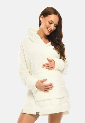Smiling pregnant woman with long brown hair wearing a white fuzzy hooded dress, gently cradling her belly with both hands.