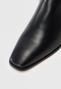 Bota de cuero negro hasta el tobillo con punta afilada, textura suave y un diseño elegante. Presenta un brillo sutil y una base delgada.