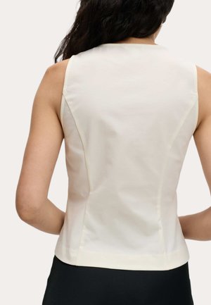 Vrouw met lang zwart haar in een mouwloos witte top en zwarte broek, gezien van achteren tegen een effen achtergrond.