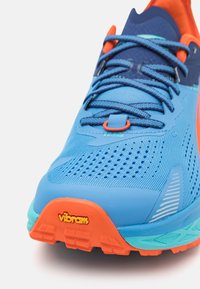 Scarpa sportiva blu con parte superiore in mesh, dettagli arancioni e suola Vibram. Caratteristiche includono perforazioni per la traspirabilità e dettagli testurizzati.