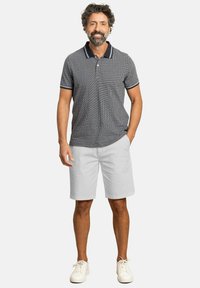Camisa polo de patrón gris con cuello azul marino, combinada con pantalones cortos blancos. Terminada con zapatillas blancas, el modelo posa con las manos en los bolsillos.