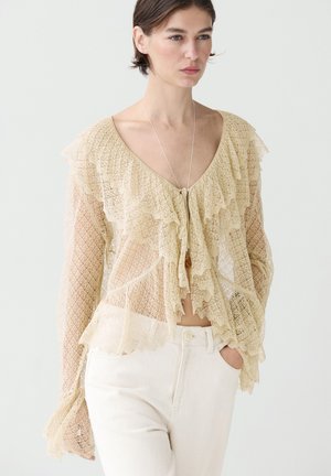 Frau trägt beige Spitzenbluse mit Rüschen, tiefem V-Ausschnitt und langen Ärmeln, kombiniert mit hochtaillierten weißen Hosen, Hand in der Tasche.