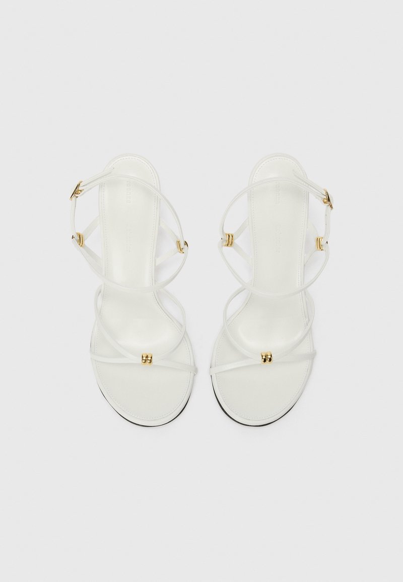 White strappy sandals with a smooth leather finish, featuring gold-tone accents and thin straps. Minimalist design with a flat sole.