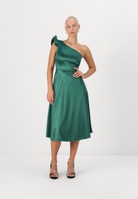 Robe en satin vert avec une épaule unique et des accents froncés. Silhouette en A avec une longueur au genou. Mannequin portant des talons noirs avec des lanières.