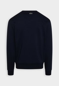 Mornarsko modra majica s dolgimi rokavi, okroglim izrezom, rebrastimi manšetami in robom ter majhnim logotipom Lacoste na zadašnji strani ovratnika.