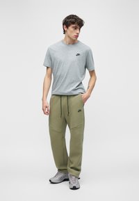 Νέος άνδρας στέκεται με το ένα χέρι στην τσέπη, φοράει γκρι μπλουζάκι Nike, χακί πράσινη φόρμα Nike και γκρι αθλητικά παπούτσια Nike σε απλό φόντο.