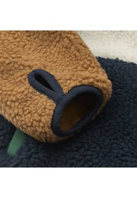 Fleeces izstrādājums ar brūnu piedurkni, kas rotāta ar tumši zilu apmali un aplītu izgriezumu. Fona ietvaros ir teksturētas tumši zilas un krēmkrāsas materiāla daļas.