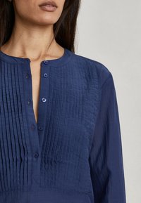 Blusa blu navy con bottoni, caratterizzata da pieghe verticali, colletto arrotondato e un tessuto liscio con semplici dettagli dei bottoni.