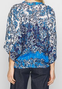 Bakifrån av en person som bär en blå och vit blommönstrad blus med vida ärmar och mörkblå byxor.