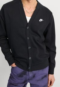 Cárdigan negro hecho de material acanalado, con cuello en V, seis botones y un pequeño logo blanco de Nike en el pecho. Combinado con pantalones morados.