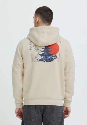 Man wearing beige hoodie with Japanese pagoda, flying cranes, red sun, and text "still water runs deep" on the back.