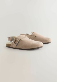 Sabots en daim beige avec une boucle réglable, un design ouvert à l'arrière, une surface texturée et une semelle extérieure en caoutchouc léger.