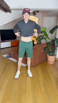 Sweatshirt zippé gris, shorts verts et baskets blanches. Cadre intérieur avec un sol en bois et une plante en pot à proximité.