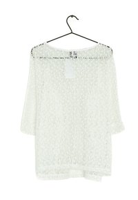 Hvid, gennemsigtig bluse med en løs pasform, 3/4-ærmer og et tekstureret mønster. Hænger på en sort krog, med en label synlig bagpå.