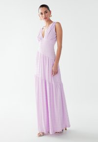 Flowy lavender maxi dress with a deep V-neck and fitted bodice. Textured fabric, tiered design, and wide shoulder straps. Open back.