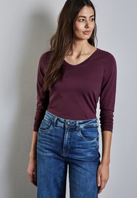 Bordeauxfarbenes Langarmoberteil mit V-Ausschnitt, kombiniert mit blauen Denim-Jeans im klassischen Schnitt und subtiler Ausbleichung des Stoffes.