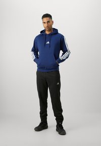 Felpa navy con strisce bianche lungo le maniche e logo sul petto, abbinata a pantaloni neri con logo, indossata con sneakers nere.