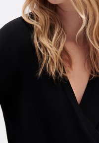 Zwarte gebreide top met een diepe V-hals en een relaxte pasvorm, met een soepele textuur en subtiele draping. Haar gestyled in losse, golvende lagen.