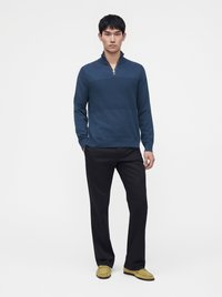 Jeune homme debout portant un pull à fermeture éclair quart bleu, un pantalon noir et des mocassins vert olive sur un fond blanc uni.