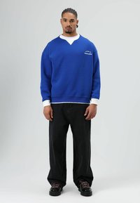 Blauwe trui met een V-hals en witte accenten, gecombineerd met zwarte broek met wijde pijpen en zwarte schoenen met roze veters.