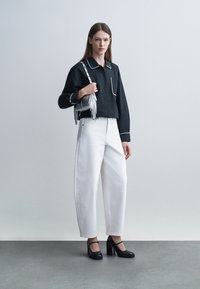 Mujer vestida con camisa negra de manga larga con ribete blanco, pantalones blancos de pierna ancha y cintura alta, tacones cuadrados negros y sosteniendo un bolso plateado con flecos.