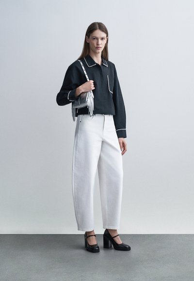Γυναίκα που φοράει μαύρο μακρυμάνικο πουκάμισο με λευκή διακόσμηση, λευκό παντελόνι φαρδύ με ψηλή μέση, μαύρα παπούτσια με τακούνι και κρατάει μια ασημένια τσάντα με κρόσια.