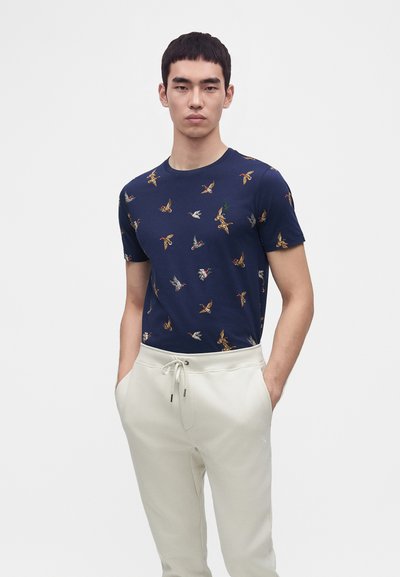 Νέος άνδρας με μπλε σκούρο t-shirt με πολύχρωμα σχέδια πουλιών και ανοιχτό μπεζ παντελόνι με κορδόνι, στέκεται με τα χέρια στις τσέπες μπροστά σε λευκό φόντο.
