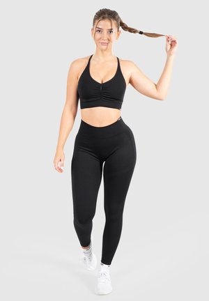 Zwarte sportbeha met geplooid ontwerp gecombineerd met hoge zwarte leggings. Model draagt witte sneakers en houdt een vlecht vast.
