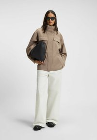 Giacca oversize color tan con due tasche frontali, abbinata a pantaloni bianchi a gamba larga e una borsa nera a forma triangolare, completata con scarpe nere.